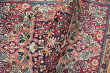 Rolled antique Kerman rug showcasing cochineal red blossoms and vines on a sand field.