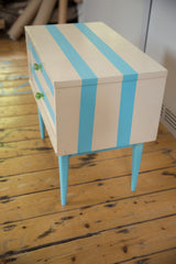Angled profile of a vintage nightstand with aqua stripes, tapered legs, and green knobs.