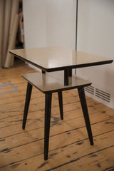 The two-tiered design of a vintage end table with light-colored tops and contrasting black tapered legs.