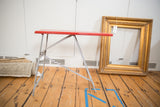 Side view of vintage children's ironing board with red top and folding metal legs.