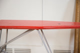 Side profile detailing the vintage wear on the red metal top and grey legs of the ironing board.