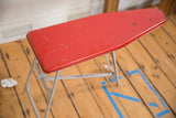 Angled view of vintage red toy ironing board with weathered gray folding metal legs.