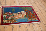 Angled view of a vintage Persian tableau rug depicting a scenic village and river.