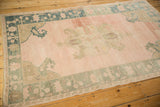 Angled view of a vintage Oushak rug with a distressed pale rose field and dusty teal borders.