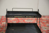 Close-up of the gold slogan 'Dependability Reliability' on the vintage metal stand.