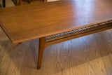 Detail view of the teak coffee table's tapered leg, wood grain, and rattan shelf.