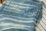 Hand-woven indigo textile with intricate ivory patterns and textured, fringed edge.