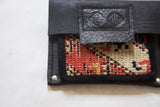 Black leather coin purse flap with embossed geometric motifs and red, cream rug fragment.