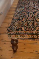 Vintage Fereghan rug ottoman corner with intricate navy, ivory, saffron pattern and carved wooden leg.