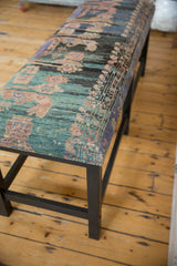 Angled view of the vintage rug bench, showing the textured upholstery and dark-stained wooden leg.