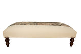 Profile view of the cream canvas ottoman with its vintage rug top and dark legs.