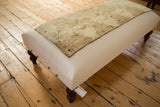 Corner view of a canvas ottoman showing its turned wooden legs and textured vintage rug top.