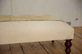 The canvas ottoman's corner construction with a dark turned wooden leg and textured rug top.