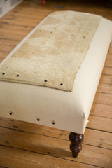 Handmade canvas ottoman showing the tan rug fragment top and a dark brown turned wooden leg.