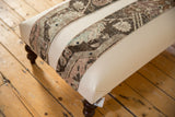 Corner view of the canvas ottoman showing a dark wood leg and snapped-on vintage rug fragment.