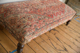 Vintage rug ottoman corner, textured rusty red and ivory pattern, visible dark carved leg.