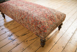 Corner view of vintage Persian rug ottoman, highlighting worn fragment and turned wooden leg.