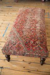 Dark carved wooden leg supports the vintage Persian rug ottoman with patterned red border.