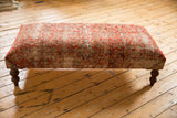 Textured vintage Persian rug ottoman featuring worn red patterns and dark wooden legs.
