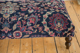 Corner of vintage Yezd rug ottoman, featuring faded navy, magenta, ivory hues and a dark wooden leg.
