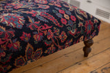 Corner of a vintage rug ottoman with navy, magenta, and blue floral Yezd textile and a wooden leg.