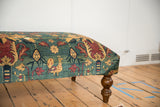 A pictorial chicken motif on an antique rug ottoman beside a curved wooden leg.