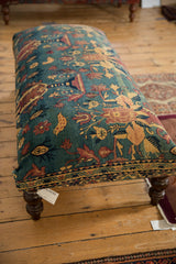 Side profile of the antique Persian rug ottoman, showing the upholstered pattern and a dark turned wooden leg.