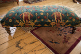 Antique rug ottoman coffee table with floral and bird motifs on a deep teal field, seen from a side angle.