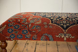 Corner detail of an antique Persian rug ottoman in cochineal red with dark navy blue floral motifs.