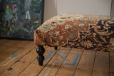 Detail of a vintage Mahal rug ottoman's corner showing the textured terracotta fabric and black turned leg.