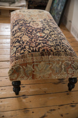 Angled view of a vintage rug ottoman, focusing on the textured border and a dark wood leg.