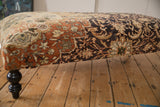 Corner detail of a vintage Mahal rug ottoman showing its worn floral upholstery and a turned black leg.
