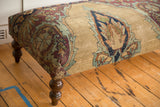 Corner of antique Kerman rug ottoman showcasing sand, aubergine, pistachio green pattern, and carved leg.