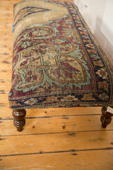 Ottoman close-up: antique Kerman rug border, sand, aubergine, dusty blue, and curved wooden leg.