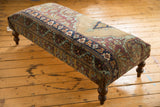 Curved corner of antique Kerman rug ottoman, highlighting pistachio, aubergine, blue, brown motifs and wooden leg.