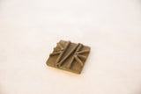 The aged patina and raised geometric design on a handmade square bronze coin.