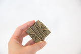 A vintage square bronze coin with a raised geometric pattern held in hand.