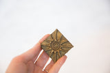 Hand-held vintage square bronze coin with spiral and radiating line pattern, aged patina.