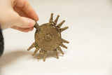 Hand-held view of aged African bronze medallion, showcasing rustic coiled pattern and radiating elements.