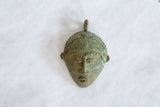 Handmade African bronze mask pendant showing textured details and a time-softened green patina.