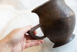 Hand gripping dark brown antique African wooden pitcher, showing its aged texture, cracks, and carved patterns.