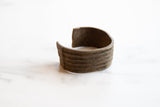 The open shape and heavily textured horizontal stripes of the antique bronze cuff.