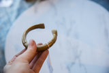 A hand holds the antique bronze cuff, showing the etched patterns and its curved form.