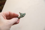 A hand holding the small bronze fish pendant, showing its green patina and spiral designs.