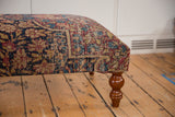 Textured antique Kerman rug upholstery on an ottoman corner beside a polished, curved wood leg.
