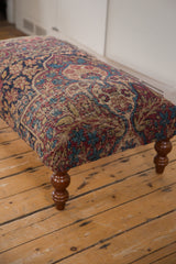Angled view of a Kerman rug ottoman showing the storied texture and a curved wooden leg.