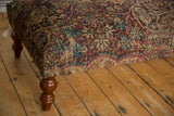 Corner view of an antique Kerman rug ottoman, detailing the worn textile and a curved leg.