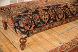 Vintage Lilihan rug ottoman corner highlighting the navy border and a turned wooden leg.