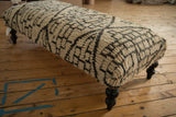 A long ottoman coffee table with plush, geometric Moroccan rug upholstery and dark wood legs.