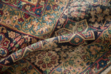 Folded antique Kerman carpet showing intricate floral pattern and pile wear in cochineal red, blue, beige.